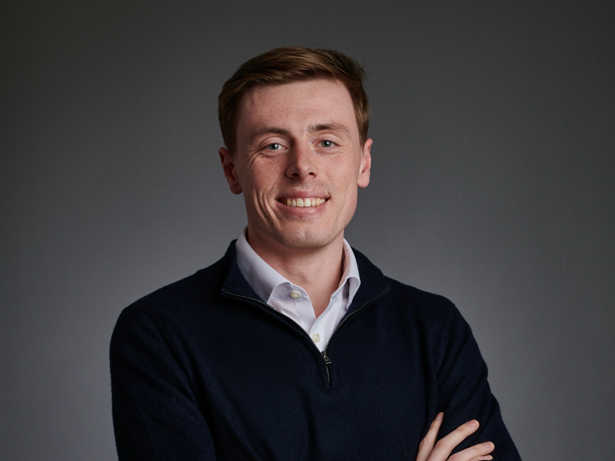 Morgan Tame smiling in a navy jumper against a grey studio background